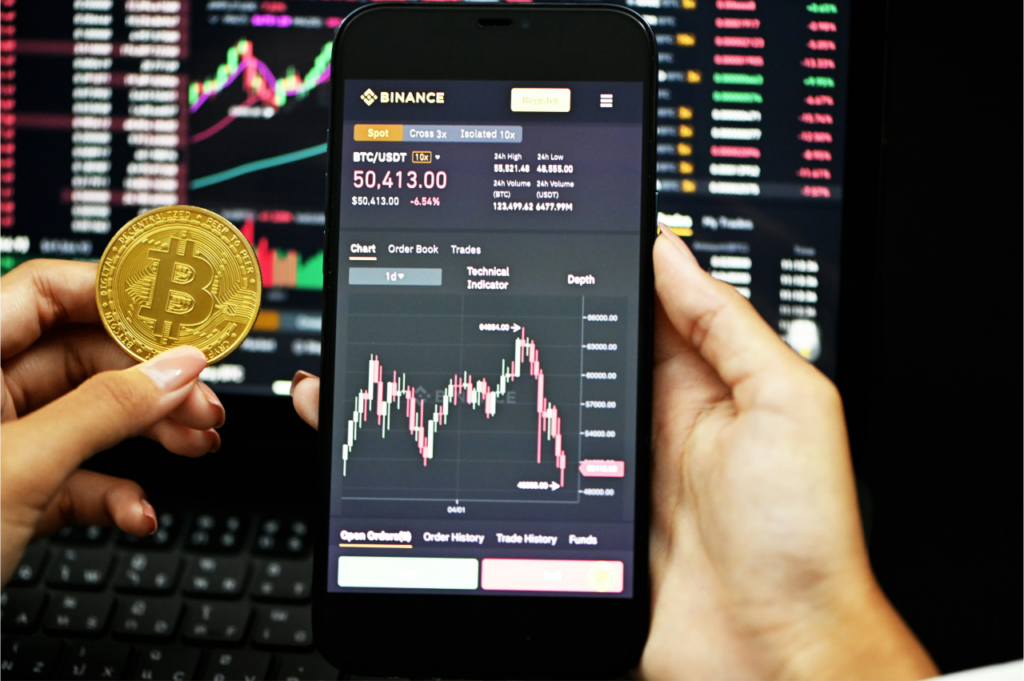 Une personne tient une pièce physique Bitcoin dans une main et un smartphone affichant l'application de trading de crypto-monnaie Binance dans l'autre, avec des graphiques et des données de marché de diverses cryptomonnaies visibles en arrière-plan.