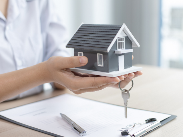 Une personne en costume tient une petite maquette d'une maison à deux étages avec un toit bleu et des murs beiges, soulignant le potentiel d'optimisation fiscale immobilière.