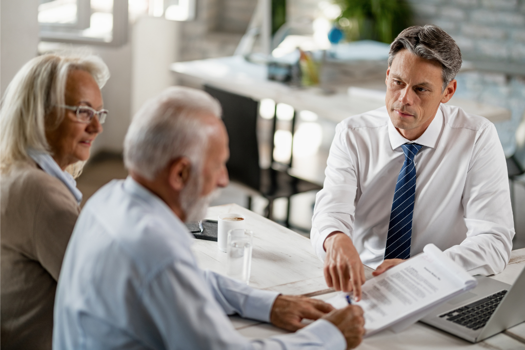 Un professionnel d'âge moyen discute de documents avec un couple de personnes âgées assis à une table dans un bureau lumineux et moderne, les aidant à planifier leur succession.