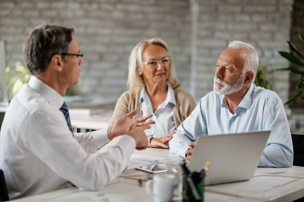 Trois personnes, deux adultes plus âgés et un adulte plus jeune, sont assis à une table dans un bureau. En pleine conversation sur le choix de leur plan de retraite, ils ont un ordinateur portable et des documents étalés devant eux.